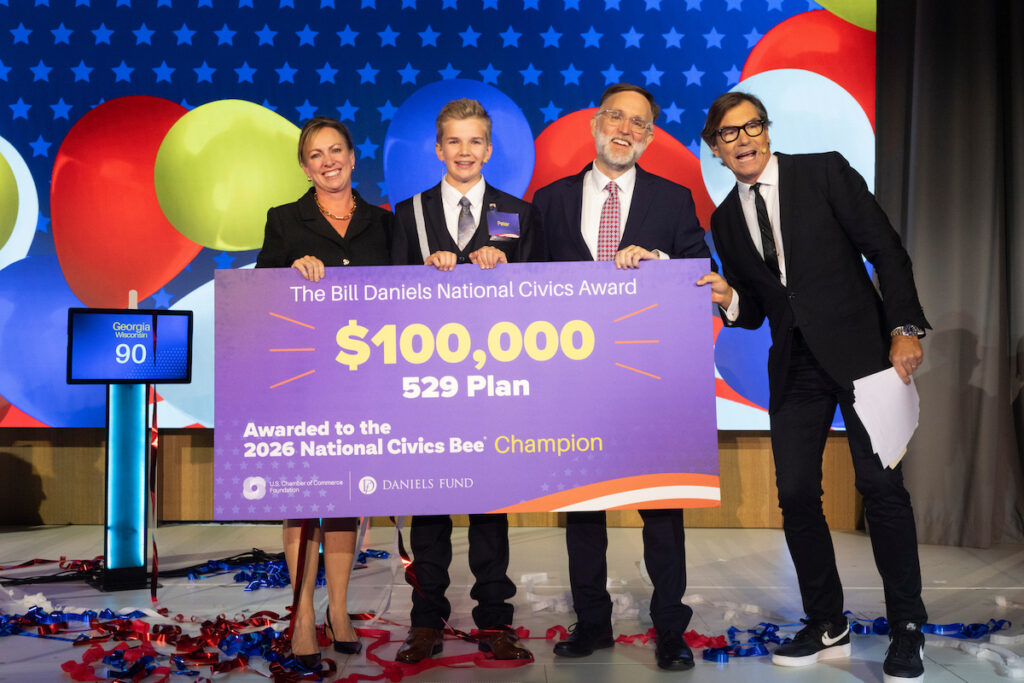 Four individuals on stage hold a large purple check for $100,000 awarded to the 529 Plan as the 2026 National Civics Bee Champion, with balloons and confetti in the background.