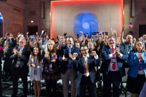 A crowd of people in formal attire, many holding American flags and smartphones, cheer and clap enthusiastically at a political event under a large illuminated backdrop.