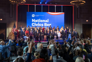 A group of young participants stands on stage at a National Civics Bee event, with an audience applauding below.