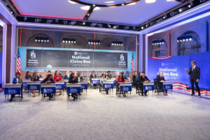 A group of young participants sits at desks during the National Civics Bee Championship, with a host standing to the side and branded signage visible throughout the studio.