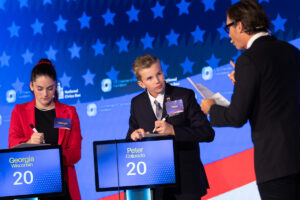 Two young participants from Georgia and Colorado, both aged 20, are seated at podiums during a debate, while a host interacts with them.